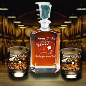May include: A glass decanter and two matching whiskey glasses with the text "Born Lucky" and "Having you as my Dad!" The decanter and glasses feature a playing card design. The decanter is filled with a brown liquid.