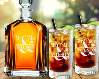 May include: A set of two personalized glasses and a decanter with the text "Mr & Mrs Wilson" etched on them. The glasses and decanter are clear glass and filled with a dark brown liquid.