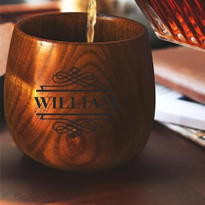 May include: A wooden cup with the name "WILLIAM" etched in black. The cup is being filled with a liquid, likely a beverage. The wood grain is visible, and the cup has a smooth, rounded shape. The overall aesthetic is rustic and elegant.