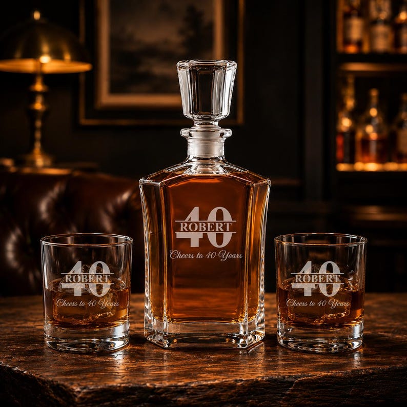May include: A crystal decanter and two matching glasses, all engraved with "ROBERT", the number "40", and "Cheers to 40 Years". The decanter is filled with amber liquid. The set is displayed on a wooden surface.