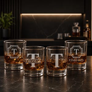 May include: Four clear whiskey glasses with ice and amber liquid. Each glass is engraved with a letter "T" and a name. The glasses are on a dark surface, with a blurred background of a bar.