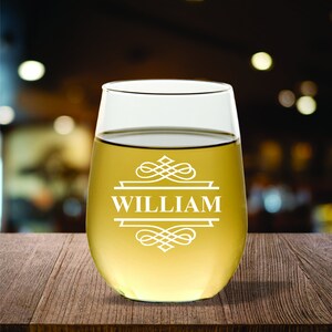 May include: A stemless wine glass filled with a light-colored liquid, possibly wine. The glass has a white decorative design with the name "WILLIAM" in the center. The glass sits on a wooden surface.