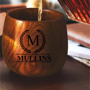 May include: A wooden tumbler with a dark stain is being filled with a liquid. The tumbler has the name "MULLINS" and the letter "M" inside a laurel wreath. The tumbler is made of wood.