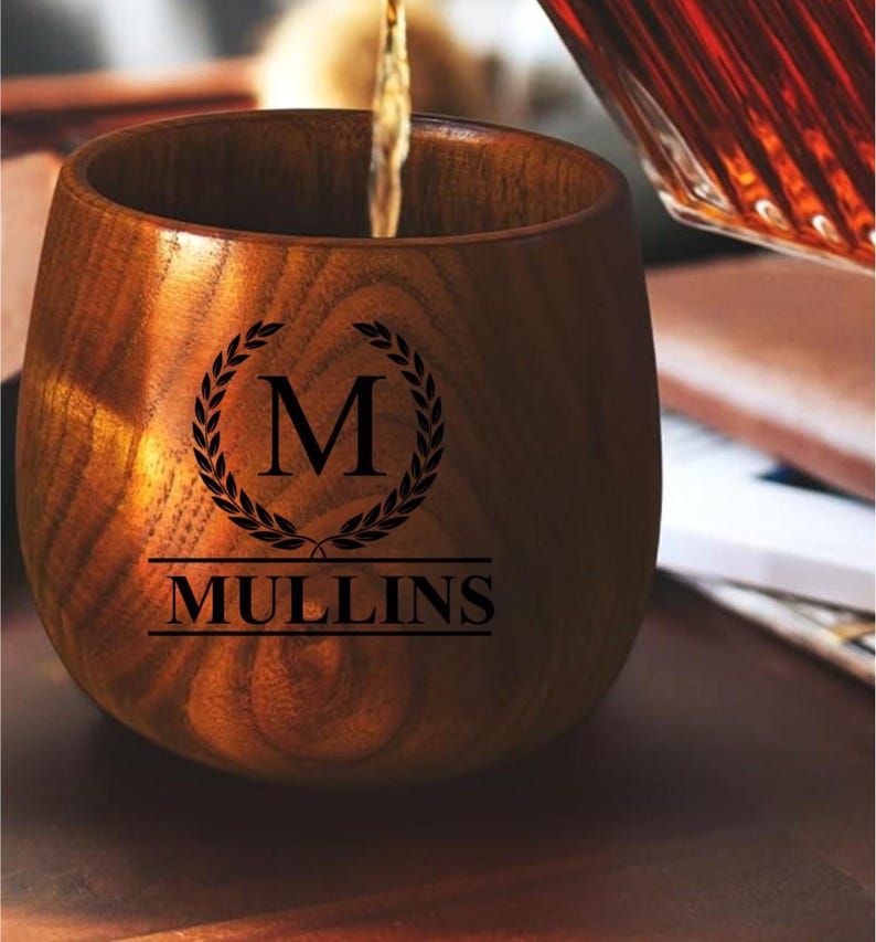 May include: A wooden tumbler with a dark stain is being filled with a liquid. The tumbler has the name "MULLINS" and the letter "M" inside a laurel wreath. The tumbler is made of wood.