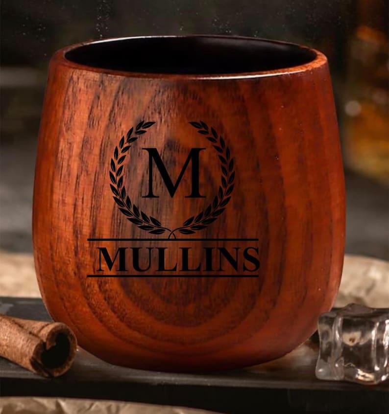 May include: A wooden tumbler with a dark brown finish. The tumbler features a black laurel wreath design with the letter "M" inside, and the name "MULLINS" below. The tumbler is designed for beverages.