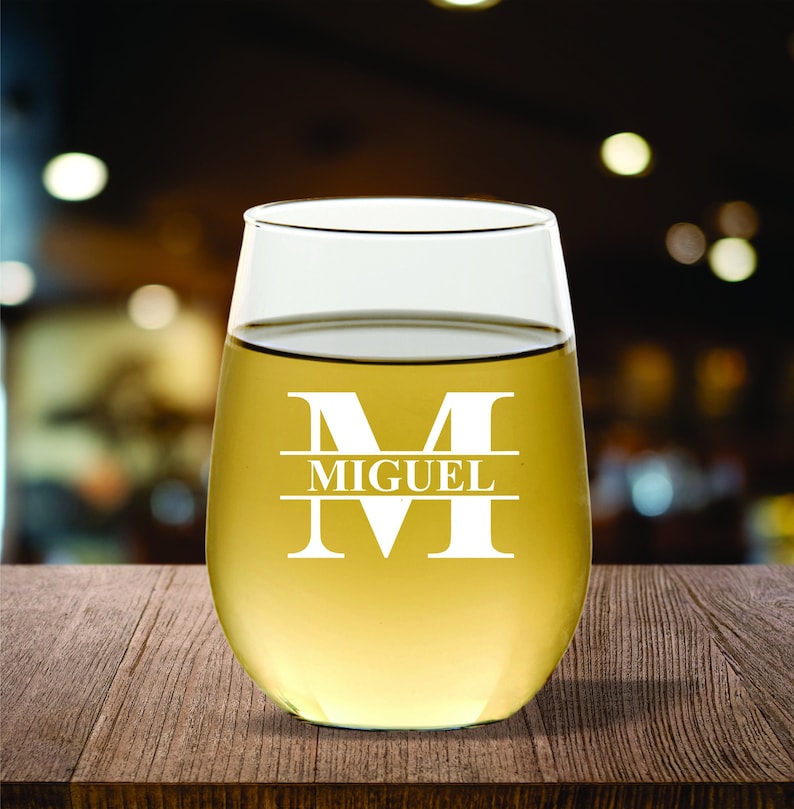 May include: A stemless wine glass filled with a light yellow liquid, likely wine. The glass has a white monogram with the name "MIGUEL" and the letter "M" etched into the glass. The glass sits on a wooden surface.