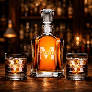 May include: A crystal whiskey decanter and two matching glasses, each engraved with the name "MARKOS" and a large initial "M". The decanter is filled with amber liquid, and the glasses contain ice.