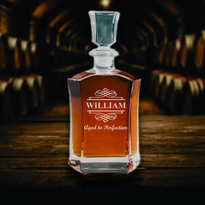 May include: A clear glass decanter filled with amber liquid, featuring a square base and a stopper. The front of the decanter is etched with the name "WILLIAM" above the phrase "Aged to Perfection" in a decorative font. The background is a dark wooden surface.