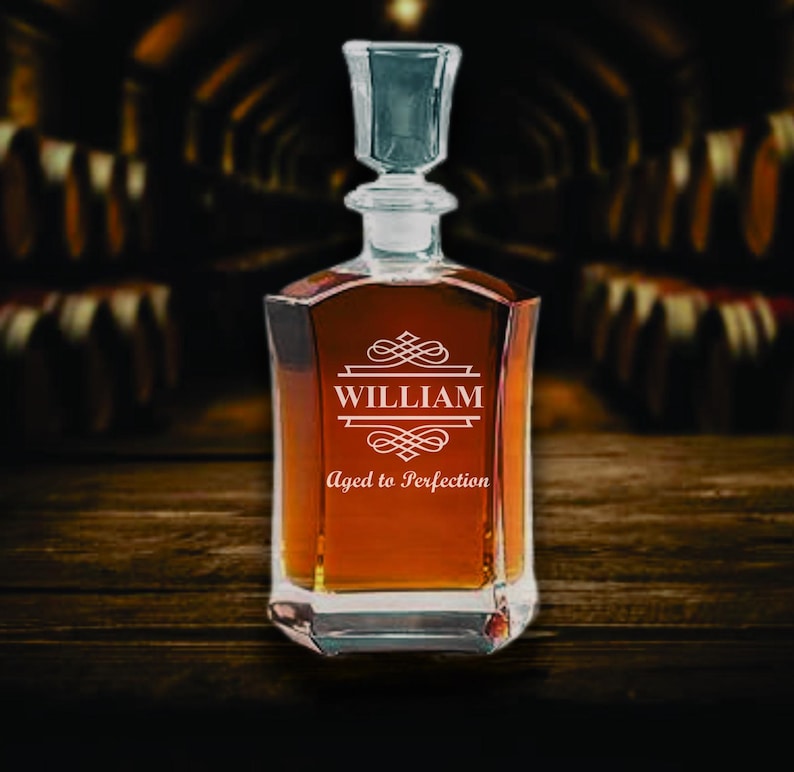 May include: A clear glass decanter filled with amber liquid, featuring a square base and a stopper. The front of the decanter is etched with the name "WILLIAM" above the phrase "Aged to Perfection" in a decorative font. The background is a dark wooden surface.