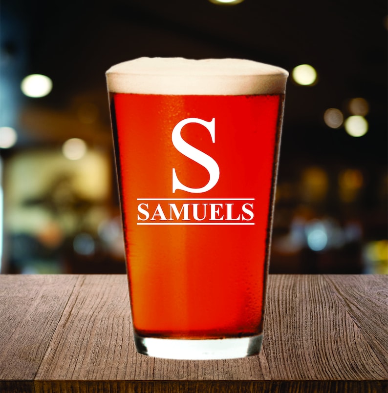 May include: A pint glass filled with a reddish-brown beverage and topped with white foam. The glass has a white "S" and the name "SAMUELS" printed on it. The glass sits on a wooden surface with a blurred background.