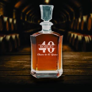 May include: A clear glass decanter filled with amber liquid, featuring a square base and a glass stopper. The front is etched with the number "40", the name "ROBERT", and the phrase "Cheers to 40 Years". The decanter sits on a wooden surface.