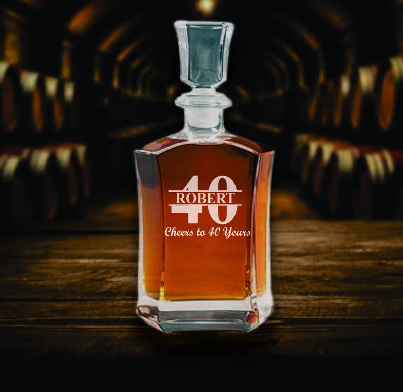 May include: A clear glass decanter filled with amber liquid, featuring a square base and a glass stopper. The front is etched with the number "40", the name "ROBERT", and the phrase "Cheers to 40 Years". The decanter sits on a wooden surface.