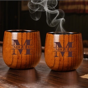 May include: Two wooden cups with a dark brown finish. Each cup has a large letter "M" and the name "MIGUEL" engraved on the front. Steam rises from the cups, suggesting they contain a hot beverage. The cups are on a wooden surface.