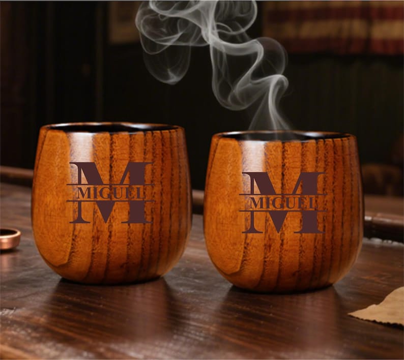 May include: Two wooden cups with a dark brown finish. Each cup has a large letter "M" and the name "MIGUEL" engraved on the front. Steam rises from the cups, suggesting they contain a hot beverage. The cups are on a wooden surface.