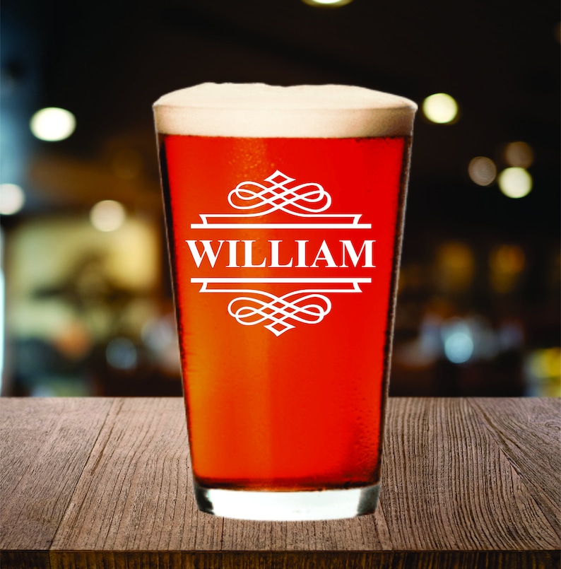 May include: A pint glass filled with a reddish-brown beverage and topped with a foamy head. The glass features the name "WILLIAM" in white, framed by decorative flourishes. The glass sits on a wooden surface.