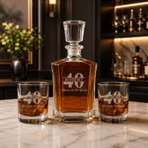 May include: A crystal decanter and two matching glasses, all engraved with "ROBERT", the number "40", and "Cheers to 40 Years". The decanter is filled with amber liquid. The set is displayed on a marble surface.