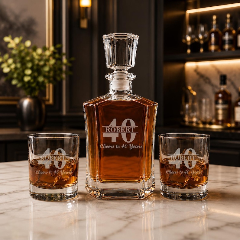 May include: A crystal decanter and two matching glasses, all engraved with "ROBERT", the number "40", and "Cheers to 40 Years". The decanter is filled with amber liquid. The set is displayed on a marble surface.