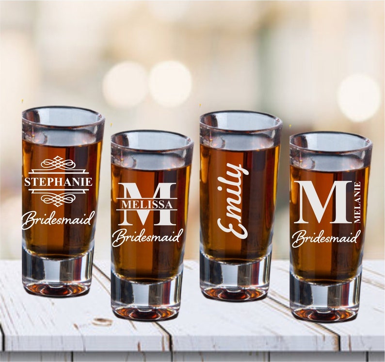 May include: Four clear glass shot glasses with personalised engravings. Each glass has a different name and the word "bridesmaid".