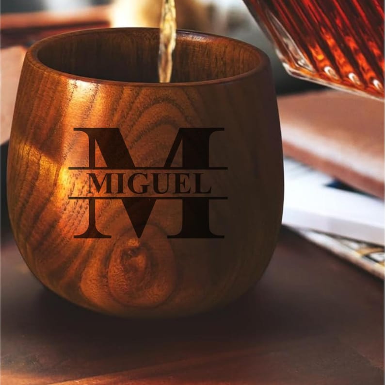 May include: A wooden cup with a dark brown finish. The cup has the name "MIGUEL" and the letter "M" engraved on the front. A liquid is being poured into the cup from a glass container.