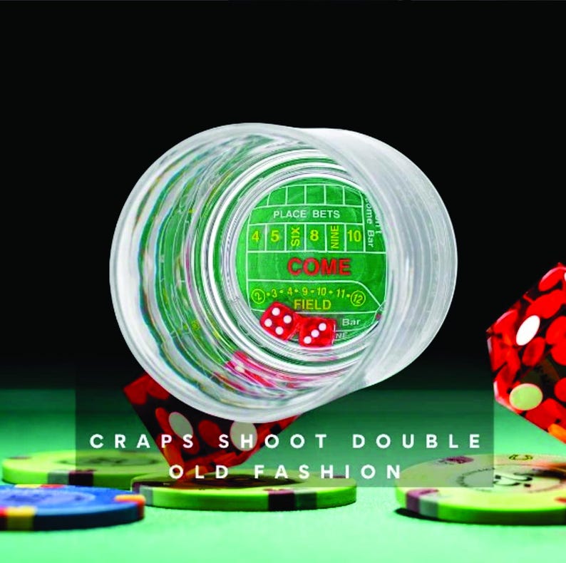 May include: A clear glass shot glass with a green felt casino craps layout inside. The glass is tipped over, revealing two red dice. The glass is surrounded by colorful casino chips.