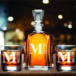 May include: A personalized whiskey decanter and two matching glasses, each with the letter "M" and the name "MILLER" etched in white. The decanter is filled with amber liquid, and the glasses contain ice and a dark beverage. The set is displayed on a wooden surface.