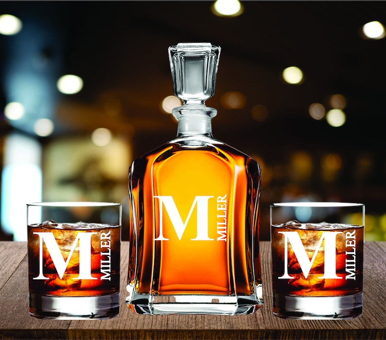 May include: A personalized whiskey decanter and two matching glasses, each with the letter "M" and the name "MILLER" etched in white. The decanter is filled with amber liquid, and the glasses contain ice and a dark beverage. The set is displayed on a wooden surface.