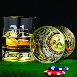 May include: Two clear glass whiskey tumblers with a gold rim. One tumbler has a roulette wheel design and the other has the name "Robert" and the number "21" etched on it. The text "Cheers to 21 Years" is also etched on the tumbler.