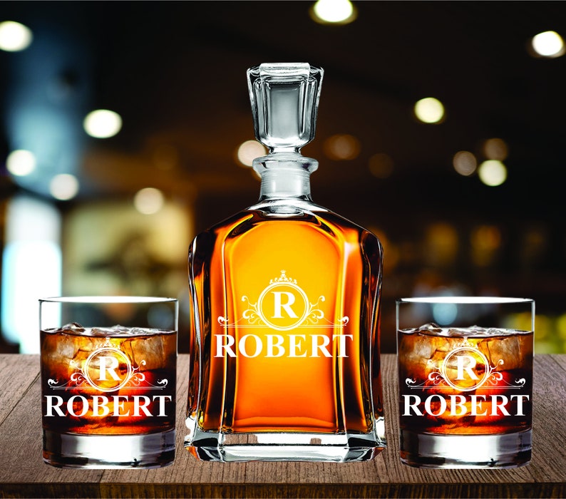 May include: A personalized whiskey decanter set with two matching glasses. The decanter and glasses are engraved with the name "ROBERT" and a monogram "R". The set is filled with amber-colored liquid and ice cubes.