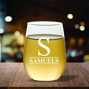 May include: A stemless wine glass filled with a light-colored liquid, possibly white wine. The glass has a large white letter "S" and the name "SAMUELS" etched on it. The glass sits on a wooden surface.
