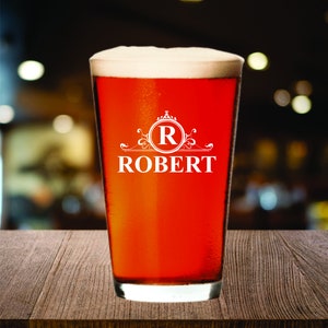 May include: A pint glass filled with a reddish-brown beverage and topped with white foam. The glass has a white design with the letter "R" inside a circle, and the name "ROBERT" below it.