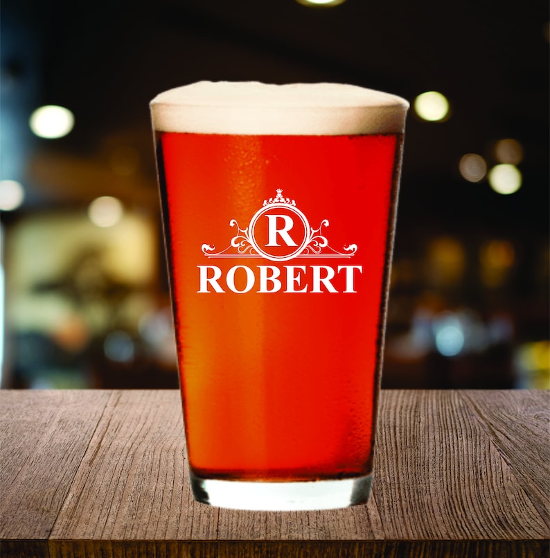 May include: A pint glass filled with a reddish-brown beverage and topped with white foam. The glass has a white design with the letter "R" inside a circle, and the name "ROBERT" below it.