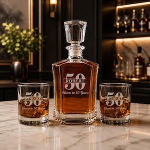 May include: A crystal decanter and two matching glasses, each engraved with "ROBERT", the number "50", and "Cheers to 50 Years". The decanter is filled with amber liquid. The set is displayed on a marble surface.