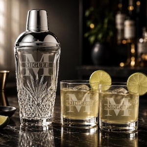 May include: A personalized cocktail shaker and two rocks glasses with the name "MIGUEL" and the initial "M" etched into the glass. The glasses contain a golden-colored liquid, ice, and lime wedges. A jigger and a small black bowl are also visible.