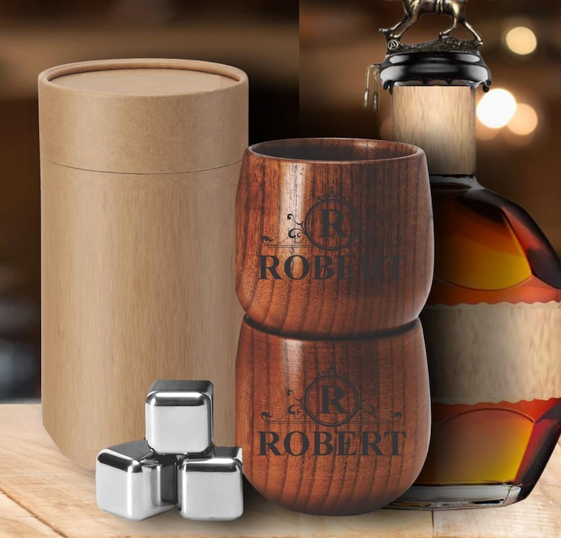 May include: A wooden tumbler set with the name "ROBERT" engraved on each cup. The set includes stainless steel ice cubes and a cylindrical cardboard container. A bottle of liquor is visible in the background.