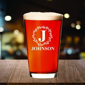 May include: A pint glass filled with a reddish-brown beverage and topped with white foam. The glass features a white floral wreath design with the letter "J" and the name "JOHNSON" in a serif font.