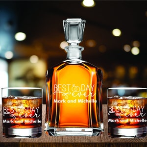 May include: A clear glass decanter and two matching glasses filled with amber liquid and ice. The decanter and glasses are engraved with the words "BEST DAY EVER" and the names "Mark and Michelle". The set is displayed on a wooden surface.