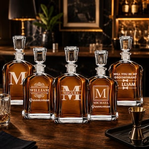 May include: Five personalized whiskey decanters with glass stoppers are displayed on a wooden bar. Each amber-colored decanter features a different initial or name, with text such as "William Groomsman" and "Will you be my Groomsman?"
