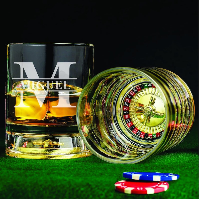 May include: Two clear glass tumblers with a gold rim. One tumbler has a personalized inscription with the letter "M" and the name "MIGUEL". The other tumbler has a roulette wheel design on the bottom. Both tumblers are sitting on a green surface with two red and blue poker chips.