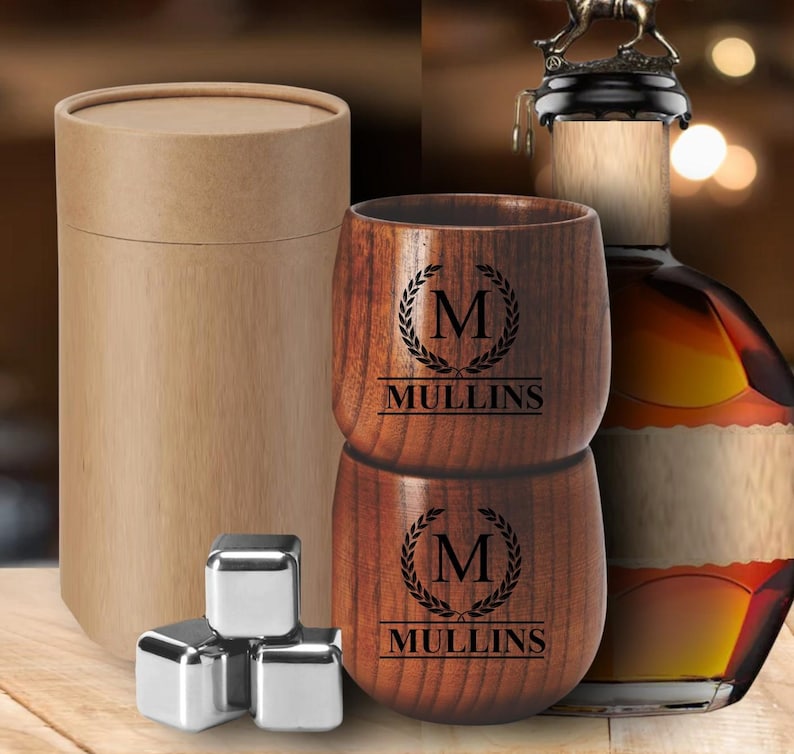 May include: A wooden tumbler set with the name "Mullins" and the letter "M" engraved on the front. The set includes a cylindrical cardboard container, stainless steel ice cubes, and a bottle of liquor with a decorative stopper.