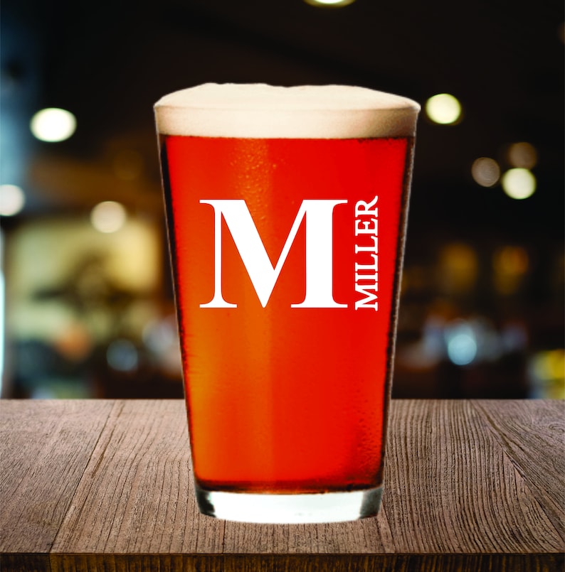 May include: A pint glass filled with a reddish-brown beverage and topped with white foam. The glass has the letter "M" and the name "MILLER" in white lettering. The glass is on a wooden surface.