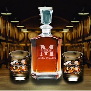 May include: A personalized decanter set with two matching glasses. The decanter and glasses feature the name "MIGUEL" and the phrase "Aged to Perfection". The decanter has a clear stopper and is filled with amber liquid.