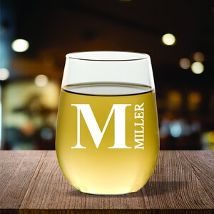 May include: A stemless wine glass filled with a light-colored liquid, possibly white wine. The glass has a large white letter "M" and the name "MILLER" printed on it. The glass sits on a wooden surface.