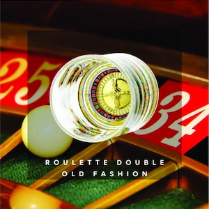May include: A close-up of a roulette wheel inside a clear glass shot glass. The roulette wheel is gold with black and red numbers. The shot glass is sitting on a wooden surface with red and white numbers.