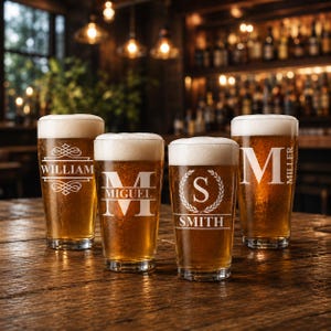 May include: Four pint glasses filled with beer and topped with white foam, each with a different personalized design. The glasses are engraved with the names "William", "Miguel", "Smith", and "Miller". The glasses are on a wooden table in a bar setting.