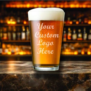 May include: A pint glass filled with beer and topped with a foamy head. The glass has the words "Your Custom Logo Here" etched on the front. The background is a blurred image of a bar.