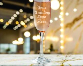 Peut inclure: Un verre à champagne rempli d'une boisson rose, avec le texte "Your Wedding Logo Here" gravé sur le verre. Le verre a une longue tige et repose sur une surface semblable à du marbre, avec un arrière-plan flou.