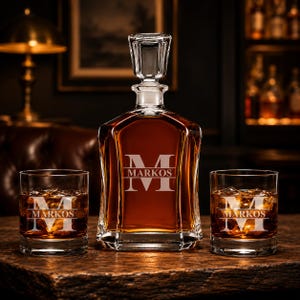 May include: A personalized whiskey decanter and two matching glasses. The decanter and glasses are engraved with the letter "M" and the name "Markos". The decanter is filled with amber liquid. The set is displayed on a wooden surface.
