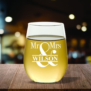May include: A stemless wine glass filled with a light-colored liquid, likely wine. The glass is etched with the words "Mr & Mrs WILSON" in a decorative font. The glass sits on a wooden surface with a blurred background.