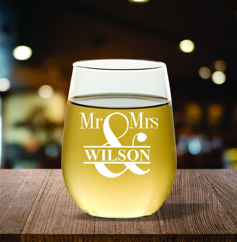 May include: A stemless wine glass filled with a light-colored liquid, likely wine. The glass is etched with the words "Mr & Mrs WILSON" in a decorative font. The glass sits on a wooden surface with a blurred background.