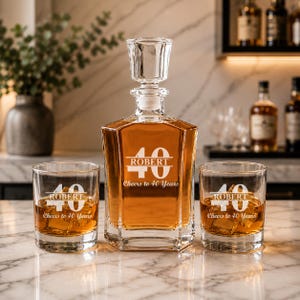 May include: A personalized whiskey decanter and two glasses with the text "ROBERT" and "40 Cheers to 40 Years" etched in white. The decanter is filled with amber liquid and has a crystal stopper. The glasses are filled with ice and amber liquid.